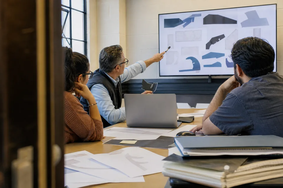 A finance team in a meeting room reviewing an ERP rollout plan on a wall screen while comparing collections vendors