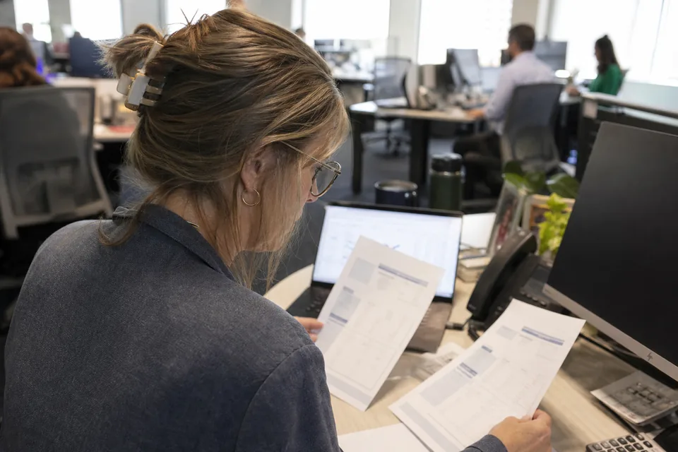 A collections manager comparing two printed AR software proposals next to a laptop with an aging report