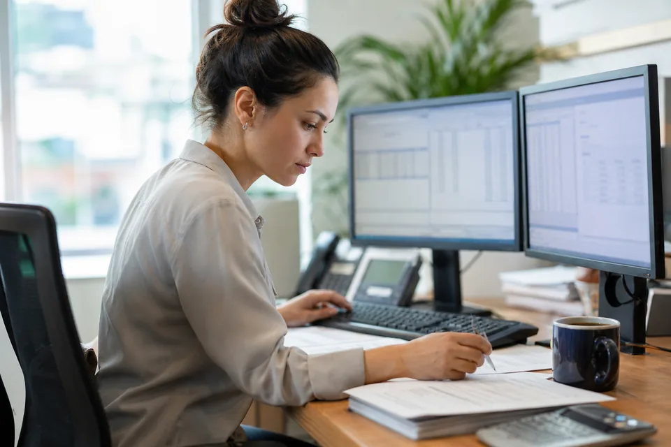 A controller at a desk with an accounting ledger on one monitor and open customer invoices on the other