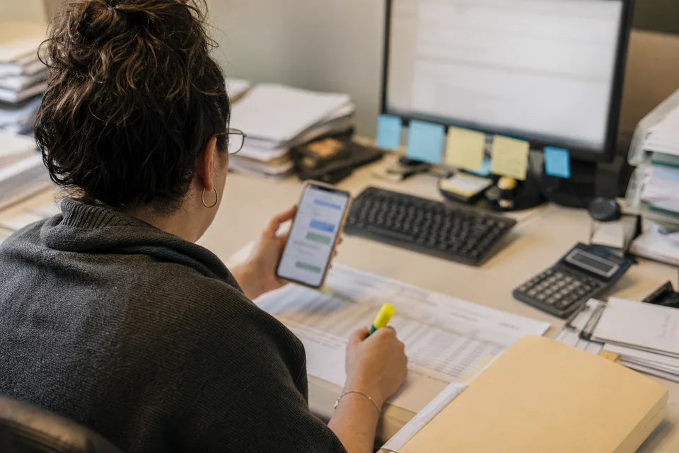 An AR specialist reviewing an aging printout while checking a customer text reply on a phone